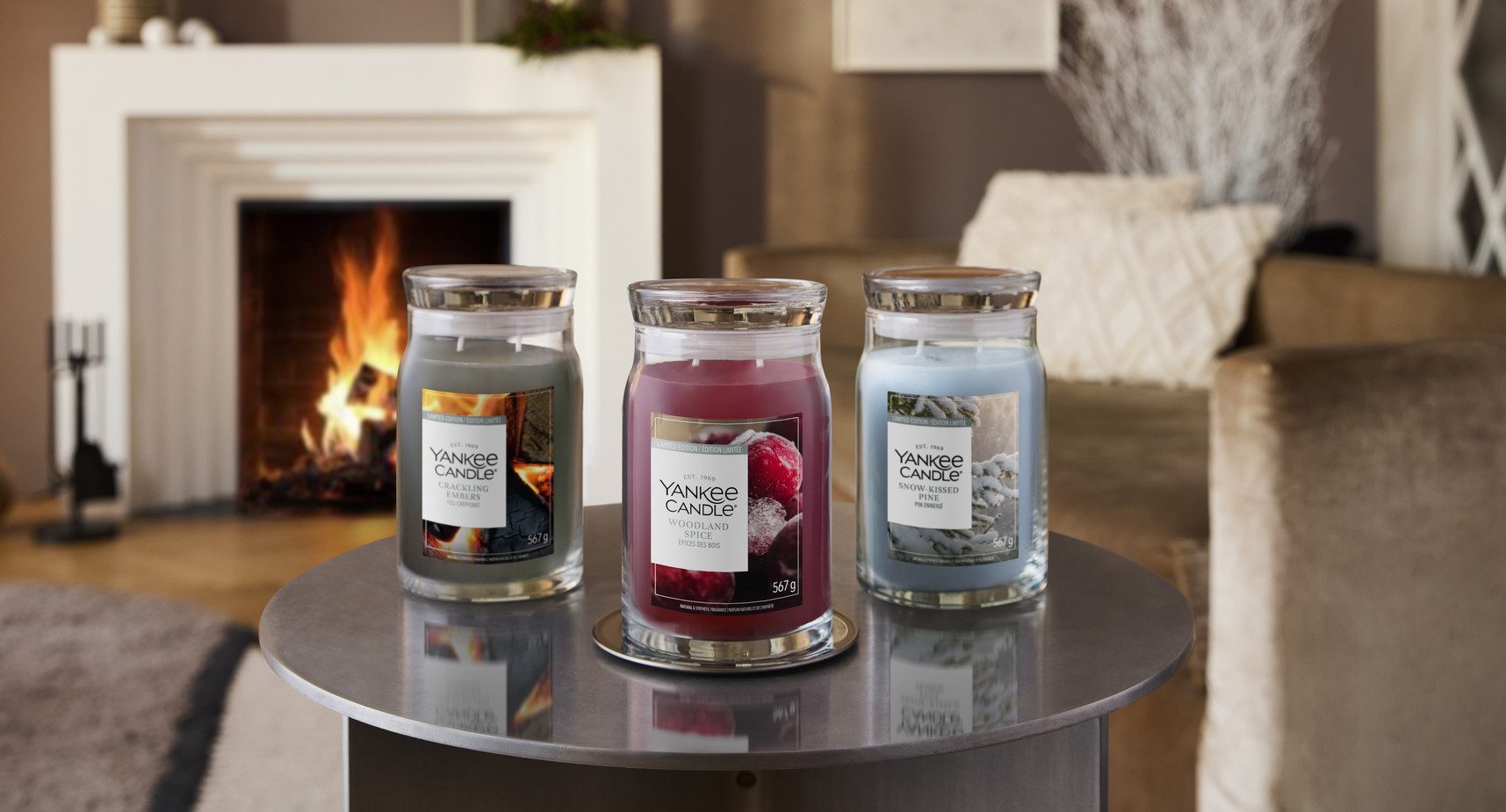 Three Yankee Candle jars on a round table in a cozy living room with a lit fireplace in the background, each labeled with its scent (Crackling Ember, Woodland Spice, Snow-Kissed Pine).